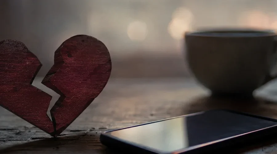 A broken heart symbol beside a smartphone and coffee cup, representing breaking up with a text message.